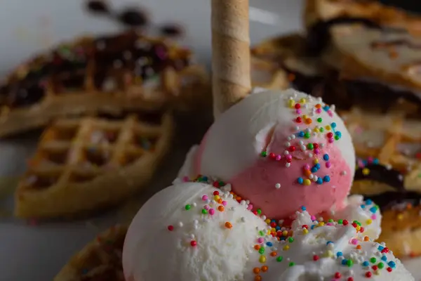 Vanilyalı ve çilekli dondurma, renkli karamel topları, waffle külahı ve arka planda waffle..