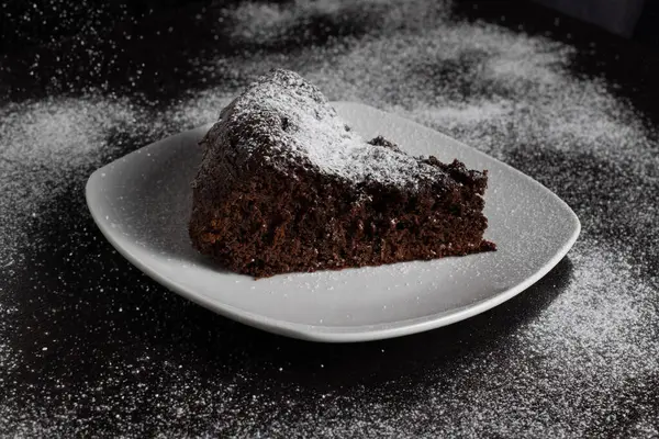 Pudra şekerli çikolatalı pasta dilimi, üzerine pudra şekeri serpilmiş bir masada servis edilir..