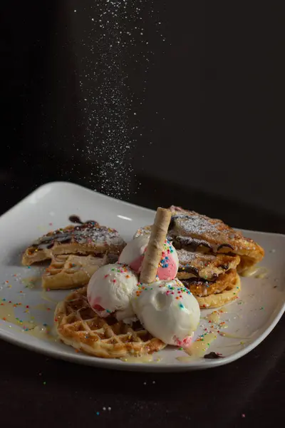 Çilekli ve vanilyalı waffleların yakın çekimi, renkli serpintiler, waffle külahı ve çikolata sosu, yukarıdan düşen pudra şekerli, koyu arkaplanlı beyaz bir tabakta servis edilir..