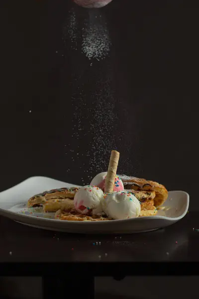 Çilekli waffle ve vanilyalı dondurma, renkli serpiştirmeler, waffle konisi ve çikolata sosu, yukarıdan düşen pudra şekerli, koyu arkaplanlı beyaz bir tabakta servis edilir..