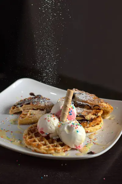 Çilekli ve vanilyalı waffleların yakın çekimi, renkli serpintiler, waffle külahı ve çikolata sosu, yukarıdan düşen pudra şekerli, koyu arkaplanlı beyaz bir tabakta servis edilir..