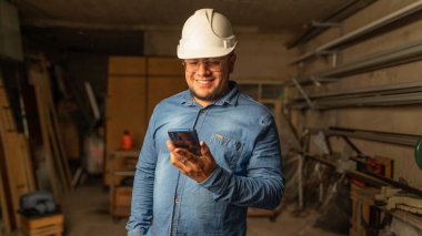 Kırsal bir metal ve ahşap atölyesinde telefonunu kontrol ederken gülümseyen bir girişimcinin yakın çekimi. Sahne, onun başarılarını yansıtan sıcak ve endüstriyel ortamda mutluluk ve başarı taşıyor..