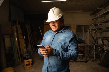 Kırsal bir metal ve ahşap atölyesinde telefonuyla konuşurken gülümseyen bir girişimcinin yakın çekimi. Görüntü bir başarı ve memnuniyet anı yakalıyor. Endüstriyel çevre atmosferi yükseltiyor.