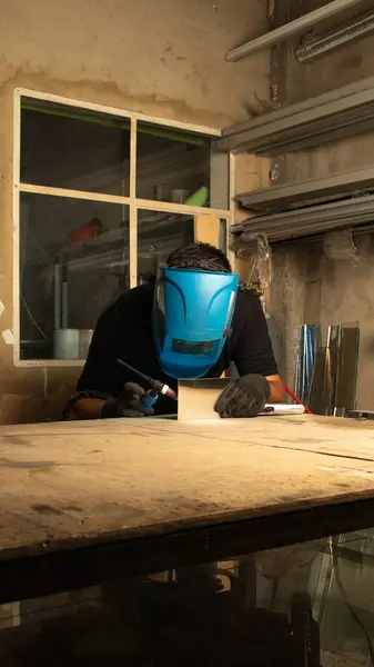 Bir kaynakçı kaynak makinesi kullanıyor, mavi koruyucu bir maske takıyor. Metal kaynaştıkça kıvılcımlar uçuşuyor. Endüstriyel bir ortamda hassasiyet ve güvenlik gösteriyorlar. Kaynak ve zanaat teması için mükemmel