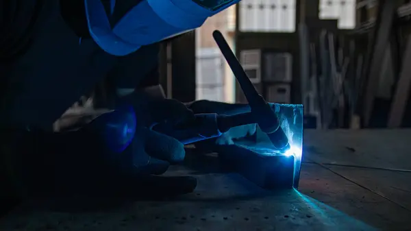 Parlak kıvılcımlar saçan kaynak makinesinin ayrıntılı görüntüsü. Mavi koruyucu bir maske kısmen görülebilir, metal işçiliğinde hassasiyet ve güvenliği vurguluyor. Endüstriyel temalar için mükemmel