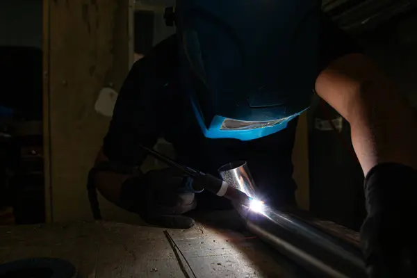 Koruyucu kasklı ve eldivenli bir işçi, bir TIG meşalesi kullanıyor, metal bir tüp kaynak yapıyor. Sahne, güvenli ve endüstriyel bir ortamda parlak kıvılcımlar ve odaklanmış işçiliği yakalıyor.