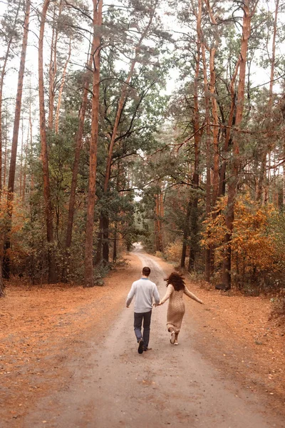 Back view on happy couple holding hands running along road in autumn forest. Young people travel in autumn in picturesque park. Yellow trees and colorful leaves. Simple pleasures of life. Blurred