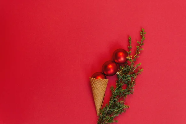 Three round toys in tasty waffle cone and next to it green branch of Christmas tree on red background. Concept of celebrating cold winter holidays in warmth. Top view, flat lay. Copy Space