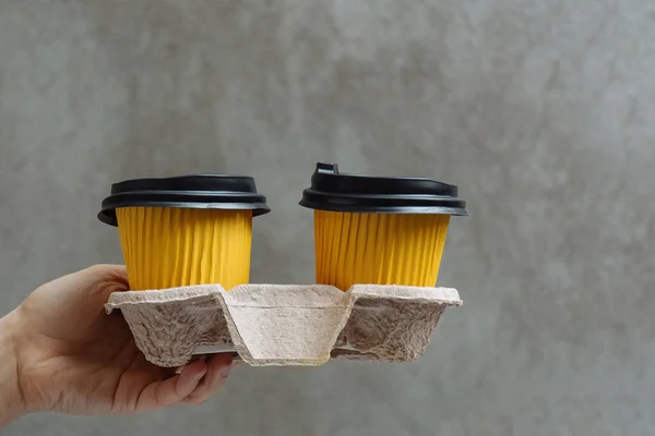 Two yellow cups with hot takeaway coffee or tea in paper holder in female hand on background of concrete wall in city. Morning pleasure before work. Copy space for text