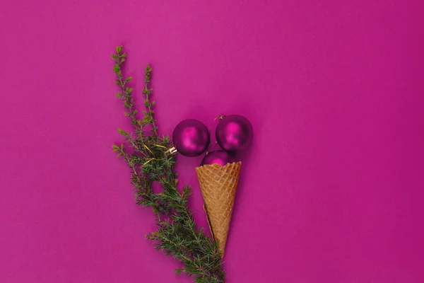 Three round, bright pink toys in waffle cone lie like delicious ice cream, and next to it is green branch of Christmas tree. Concept of celebrating cold winter holidays in warmth. Top view, flat lay