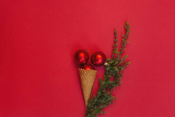 Three round toys in waffle cone lie like delicious ice cream and next to it is green branch of Christmas tree on red background. Concept of celebrating cold winter holidays in warm. Top view, flat lay