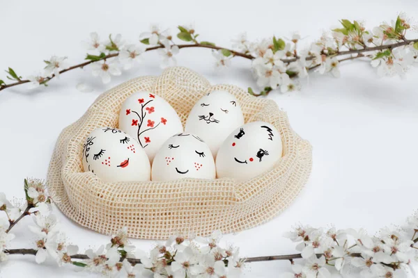 Defocused focus on foreground sprigs of flowers. Easter beautifully painted eggs in eco-friendly mesh bag on white table. Funny spades, flowers, cats face are painted on eggs. Copy space