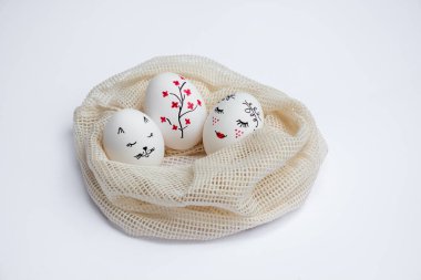 Three beautifully painted Easter eggs lie in environmentally friendly mesh bag on white table. Funny faces and twig with red flowers are painted on eggs. Copy space. Happy holidays