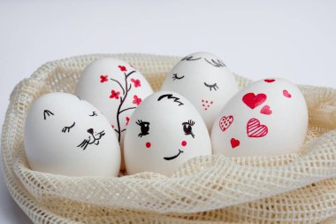 Close-up Delicately painted Easter eggs lie in environmentally friendly mesh bag on white table. Funny peaks and twig with red flowers are drawn on eggs. Copy space. Happy Easter