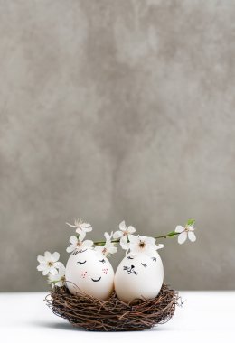 Happy Easter. White Easter eggs in wicker wooden nest on white table on gray background. View is straight. Blossoming branch of cherry with white flowers. Stylish spring template with copy space