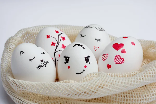 Close-up Delicately painted Easter eggs lie in environmentally friendly mesh bag on white table. Funny peaks and twig with red flowers are drawn on eggs. Copy space. Happy Easter