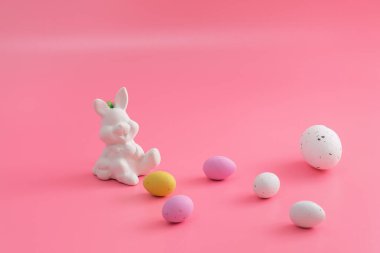Easter greeting card with white bunny and sweet colorful little eggs lying on pale pink background. Copy space for text.