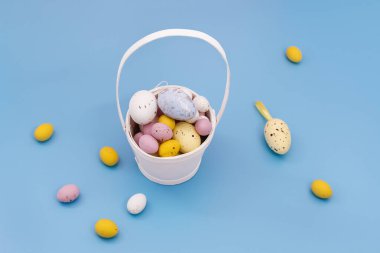 Happy Easter card. Multi Colored sweet chocolate eggs and decorative eggs in a white basket and on a blue background. Yellow, pink. Copy space for text. Flat Lay
