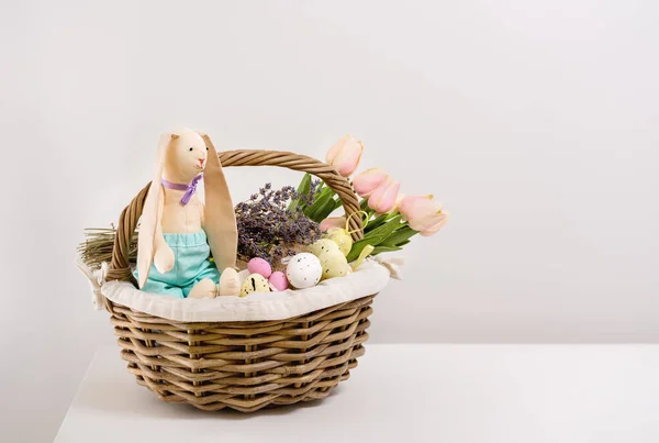 Vintage postcards for Easter, happy holidays. Preparing for picnic in nature. In basket there is bouquet of tulips and lavender, toy rabbit, sweets and Easter eggs on white table. Copy space