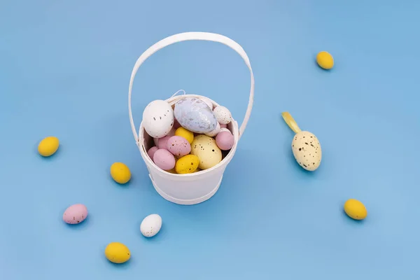 Happy Easter card. Multi Colored sweet chocolate eggs and decorative eggs in a white basket and on a blue background. Yellow, pink. Copy space for text. Flat Lay