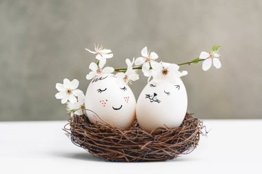 Mutlu Paskalya tebrik kartları. Hasır ahşap yuvalarda beyaz Paskalya yumurtaları gri arka plandaki beyaz masada. Nazik boyalı yüzler. Görüntü doğrudan. Beyaz çiçekli kiraz dalları. Boşluğu Kopyala