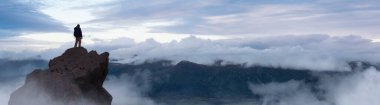 Adventurous Man Hiker standing on top of peak with rocky mountain in background. Adventure Composite. 3d Rendering rocks. Aerial Image of landscape from Yukon, Canada. Sunset Sky