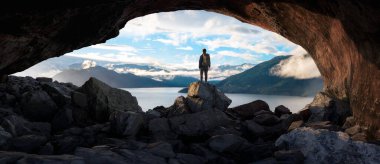 Rocky Mağarasında Duran Maceracı Adam. Arka planda dağlar ve okyanus. Gün batımı ya da gün doğumu. 3d Hazırlama