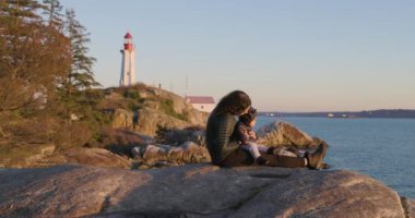 Anne ve Baby Boy okyanus kıyısında Rock 'ta oturuyorlar. Arka planda deniz feneri, Sunset. Fener Parkı, Batı Vancouver, BC, Kanada.