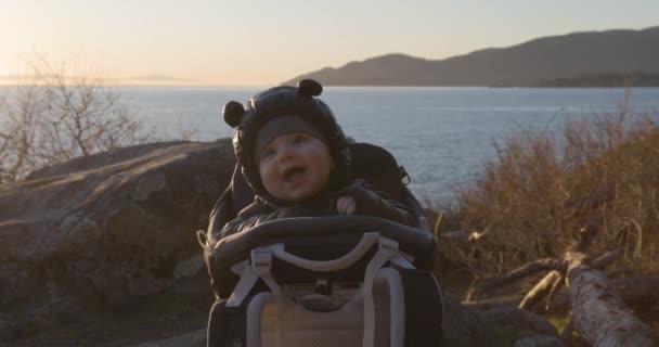 ベイビー ボーイが微笑みながら 風光明媚な海岸でロックでハイキングキャリアに座った サンセット ライトハウスパーク ウェストバンクーバー カナダ — ストック動画