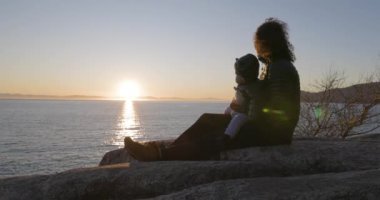 Anne ve Baby Boy okyanus kıyısında Rock 'ta oturuyorlar. Gün batımı. Fener Parkı, Batı Vancouver, BC, Kanada.