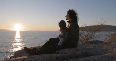 Anne ve Baby Boy okyanus kıyısında Rock 'ta oturuyorlar. Gün batımı. Fener Parkı, Batı Vancouver, BC, Kanada.