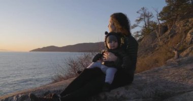 Anne ve Baby Boy okyanus kıyısında Rock 'ta oturuyorlar. Gün batımı. Fener Parkı, Batı Vancouver, BC, Kanada.
