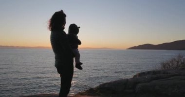 Anne, okyanus kıyısında Rock 'ın üzerinde duruyor, kucağında bebek var. Sunset 'e doğru bakıyor. Fener Parkı, Batı Vancouver, BC, Kanada.