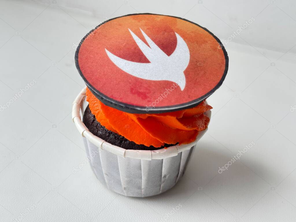 Close-up image of a chocolate cupcake with vivid orange frosting, topped with an edible decoration featuring the Swift programming language logo. Ideal for themes involving programming, tech culture, Swift development, software engineering lifestyle.