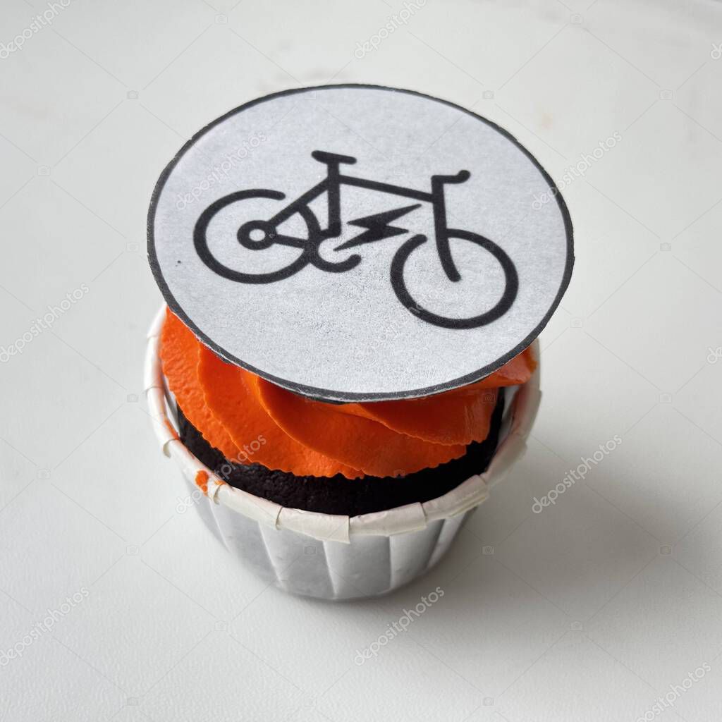 Chocolate cupcake with bright orange frosting, topped with an edible e-bike icon on white background. A creative mix of dessert and eco-mobility, perfect for themes like sustainability and modern lifestyle.