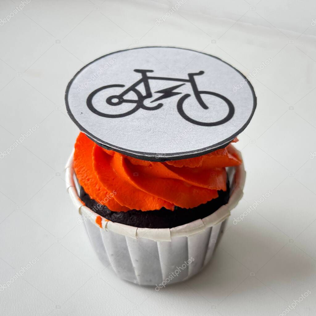 Chocolate cupcake with bright orange frosting, topped with an edible e-bike icon on white background. A creative mix of dessert and eco-mobility, perfect for themes like sustainability and modern lifestyle.