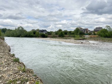 Ukrayna 'daki Bystrytsia Nadvirnianska nehrinin sakin bir bahar manzarası. Bir çakıl taşı nehir kıyısı ve sahildeki evler Yumuşak, bulutlu bir gökyüzünün altında.