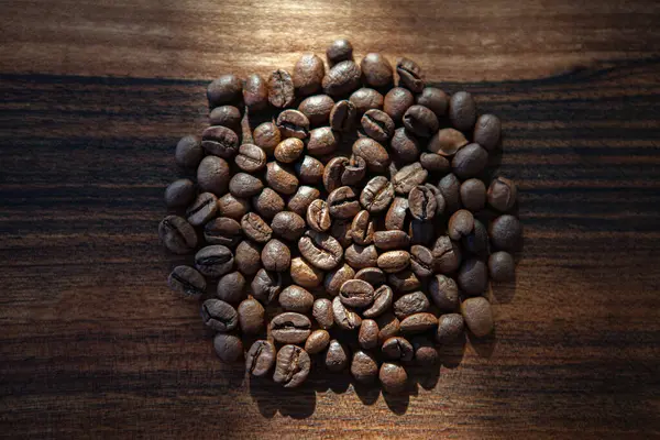 Koyu ahşap arka planda kavrulmuş kahve çekirdekleri. Kahve çekirdekleri, üzerine güneş ışığı düşen ahşap bir zemine saçılmış.