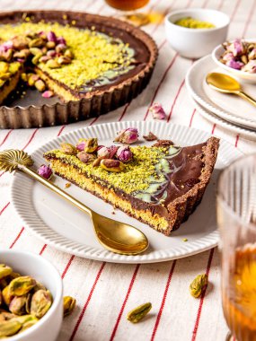 Üzerinde gül yaprakları ve yer fıstığı olan Dubai çikolatalı turta dilimi beyaz bir tabakta altın bir kaşık ve tabaklar ve bardaklarla servis edilir.