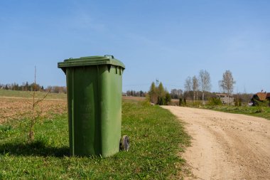 Kırsal Toprak Yolundaki Yeşil Çöp Kutusu. Güneşli bir bahar gününde kırsal bir toprak yolun kenarına yerleştirilmiş, açık arazilerle çevrili büyük yeşil çöp tenekesi..