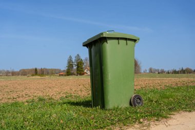 Kırsal Toprak Yolundaki Yeşil Çöp Kutusu. Güneşli bir bahar gününde kırsal bir toprak yolun kenarına yerleştirilmiş, açık arazilerle çevrili büyük yeşil çöp tenekesi..