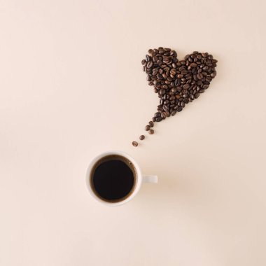 Kalp şeklinde kahve çekirdekleri, bej arka planda bir fincan siyah kahve. Aşk konsepti.