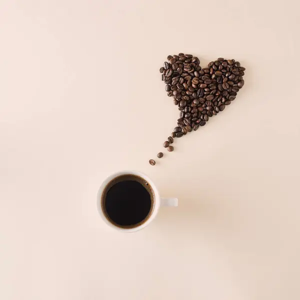 Kalp şeklinde kahve çekirdekleri, bej arka planda bir fincan siyah kahve. Aşk konsepti.