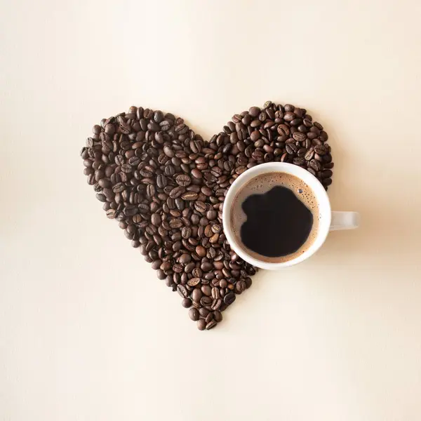 Kalp şeklinde kahve çekirdekleri, bej arka planda bir fincan siyah kahve. Aşk konsepti.
