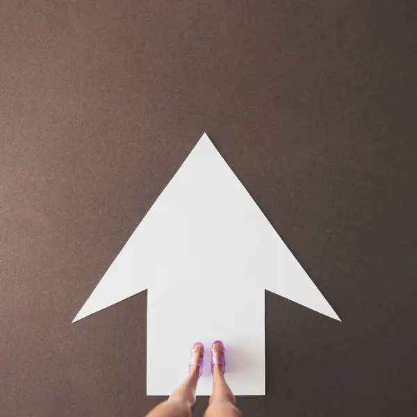 Beyaz bir ok üzerinde oyuncak bacaklı, kahverengi bir arka planda yön işareti olan minimum bileşim. Düz.