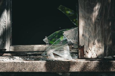 Kırık camlı bir pencere dış dünyayı yansıtır, yıpranmış ahşap bir çerçeve ve taş pervaza çarpar, çürüme ve dayanıklılık manzarası yaratır..