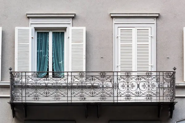 Süslü tasarımlarla işlenmiş demirden bir balkon içeren büyüleyici bir mimari detay, beyaz panjurlu iki pencere ile tamamlanmış, biri açık mavi perdeleri bir anlığına gözler önüne seren.