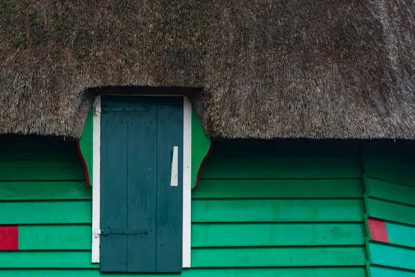 Geleneksel sazdan çatısı olan tuhaf bir bina, yatay yeşil kaplamalı bir arka fona karşı koyu mavi bir pencereye sahiptir..
