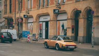 Altın sarısı bir Porsche 911, Berlin caddesinde bir New Balance mağazasının önünden geçiyor. Kum taşı binası tarihi cazibeyle modern markayı harmanlarken, cesur reklamlar ve 30 km / saat hız sınırı işareti şık bir kontrastın ve kontrollü hızın tonunu belirler..