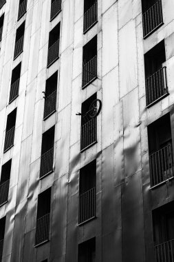 Metalik bina cephesinin siyah ve beyaz görüntüsü tekrarlanan pencere desenleri ve tuhaf bir şekilde balkona monte edilmiş bir bisiklet.